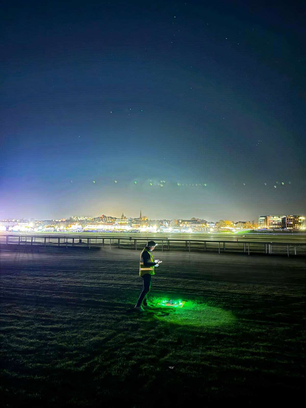 Drone operator standing on beach at night with city lights and starry sky.