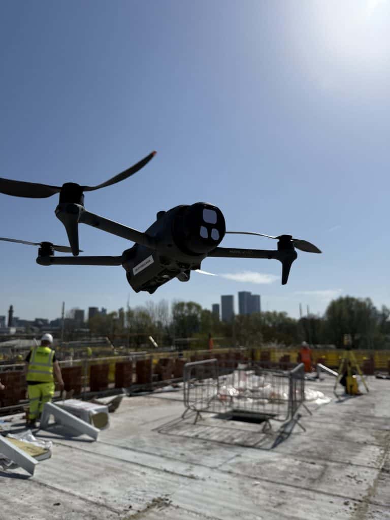 Drone flying over construction site with workers and city skyline in background.
