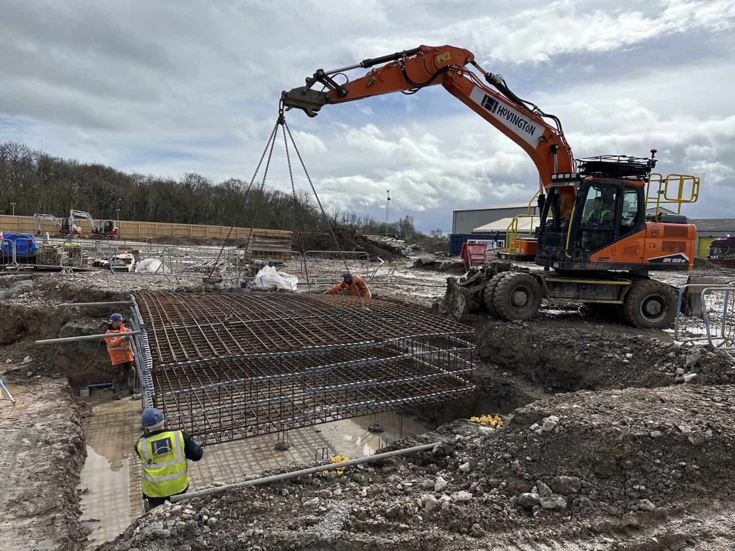 Excavator placing reinforcement bars at a construction site in cloudy weather.